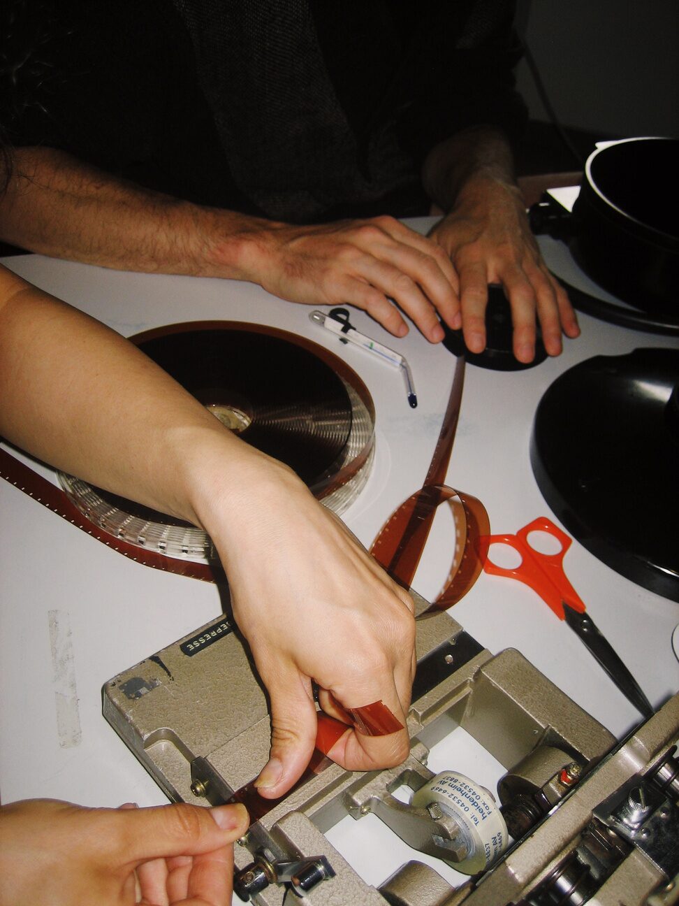 Hands cutting and splicing developed 16mm film on a film splicer beside a reel and red scissors