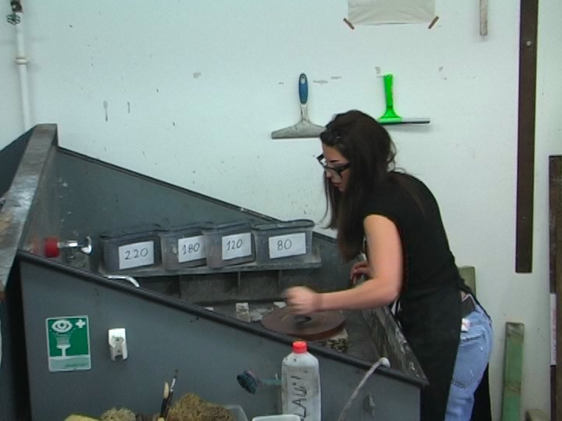 Melina Katsiri graining a lithographic stone with a levigator at the graining sink, next to boxes of abrasive grit labelled 220 to 80