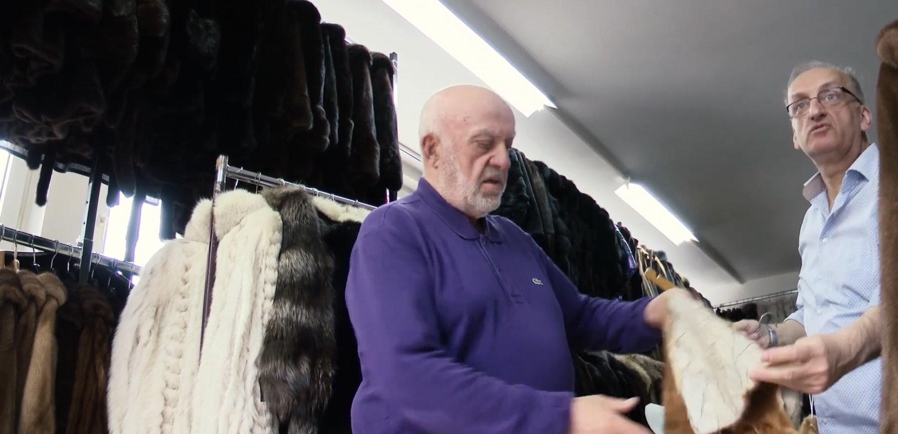 The protagonist and a colleague examining a fur between racks of coats in a fur shop